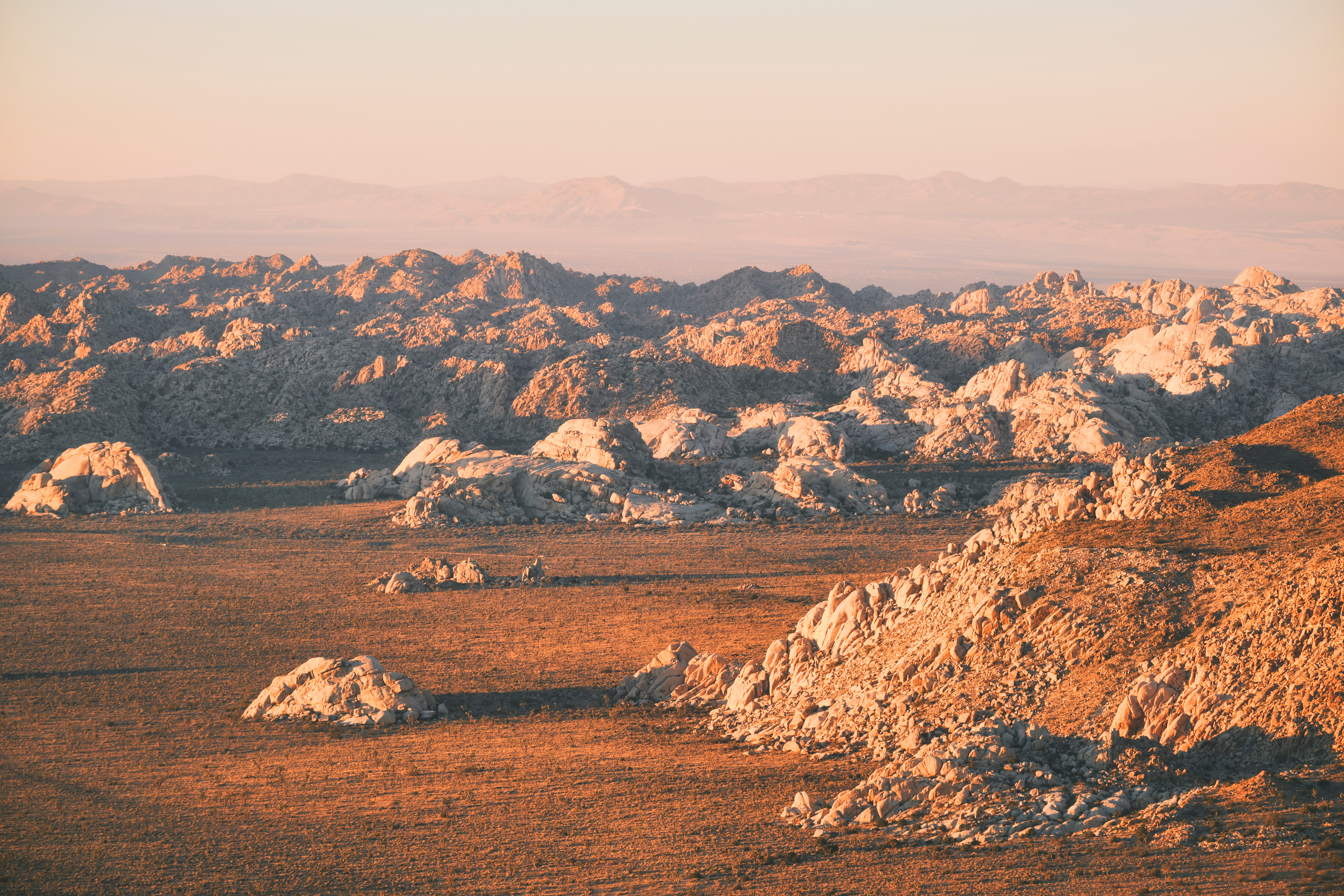 Low golden light raking across a desert basin strewn with granite boulders, hazy mountains beyond