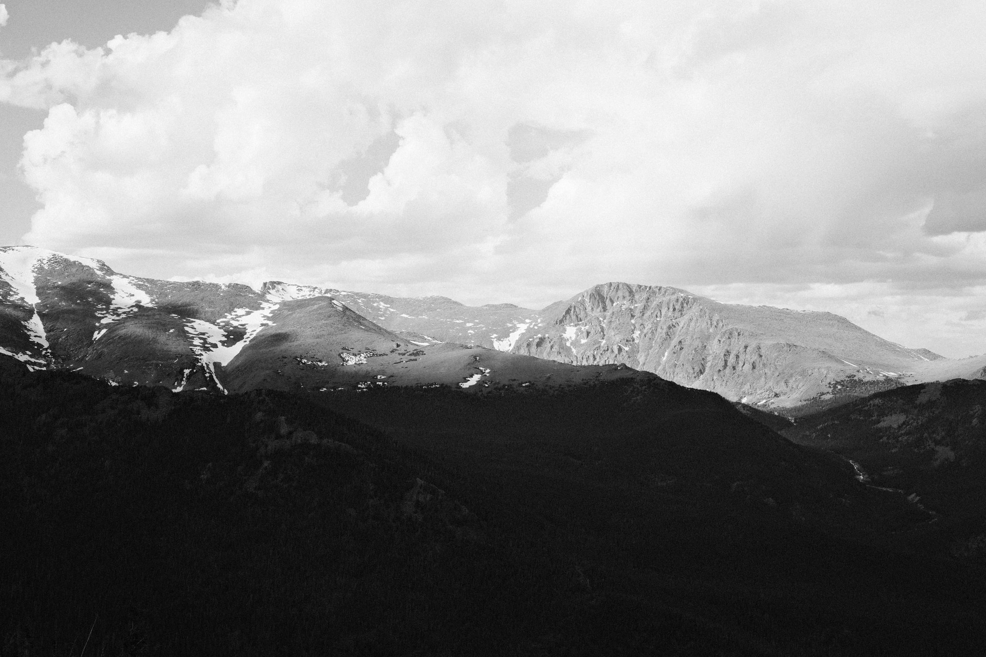 Snow-streaked peaks beneath towering clouds above dark forested ridges, in black and white