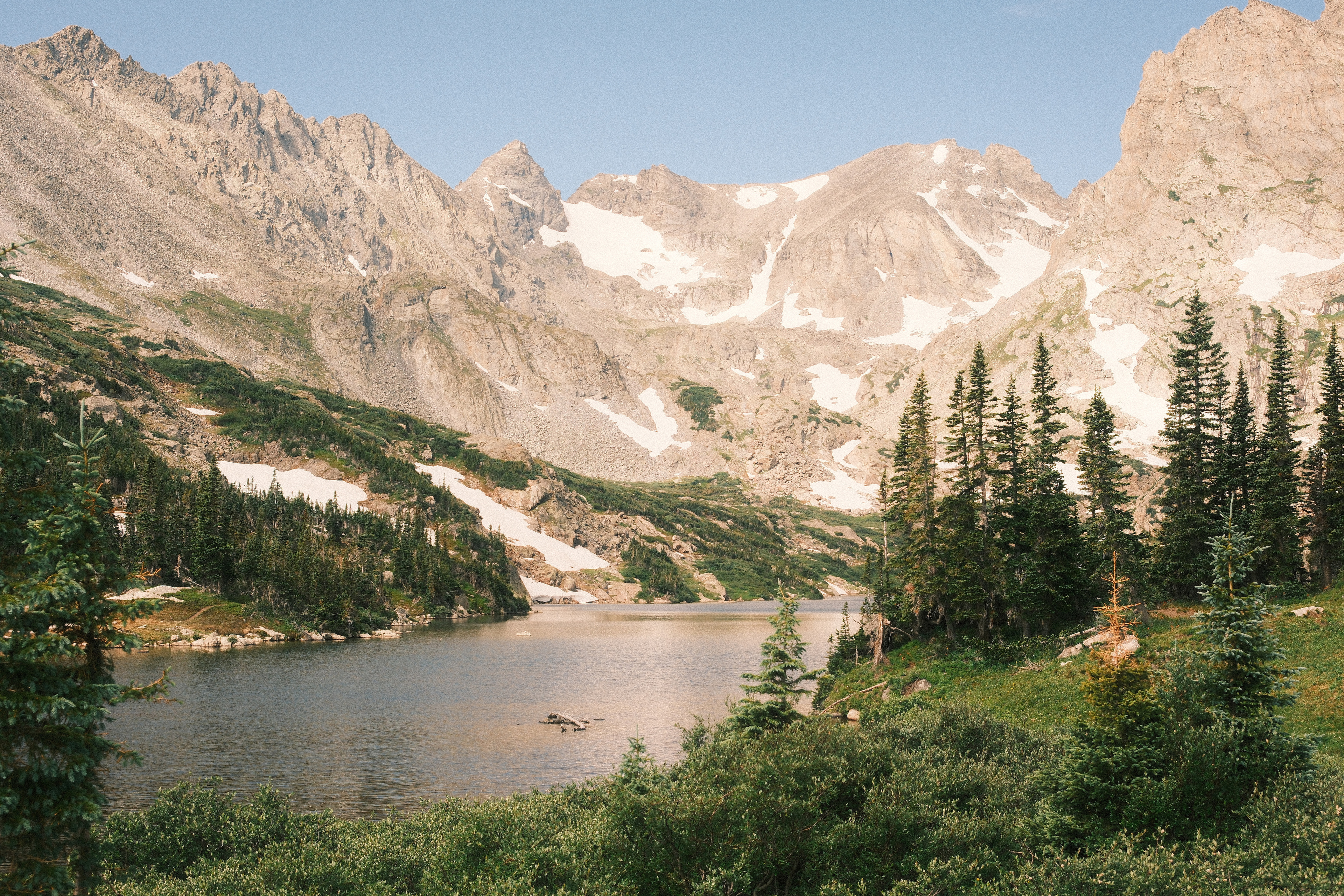 An alpine lake ringed by jagged, snow-streaked peaks with pines along the green shoreline
