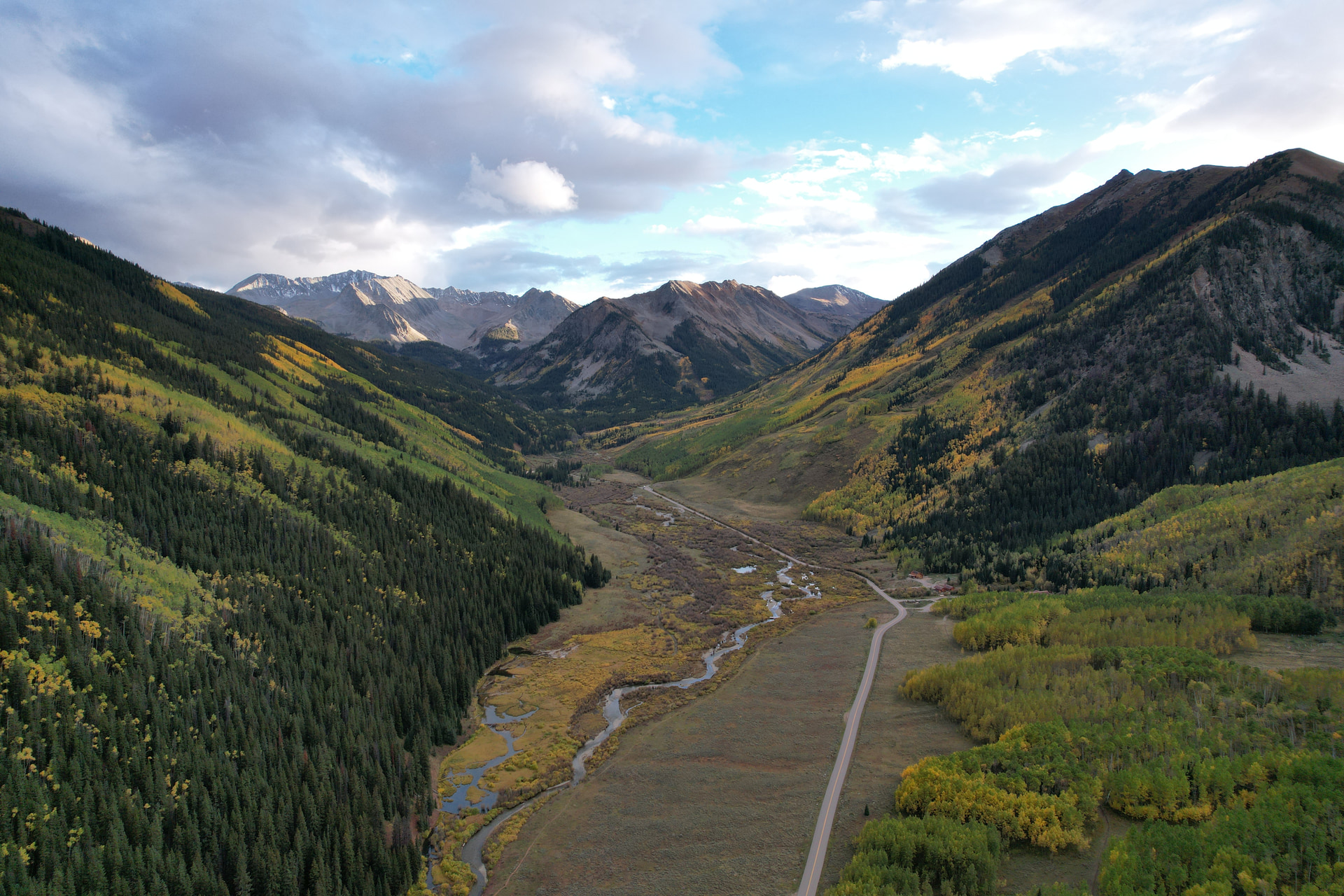 An aerial view down a mountain valley, a creek winding past golden aspens beside a dirt road
