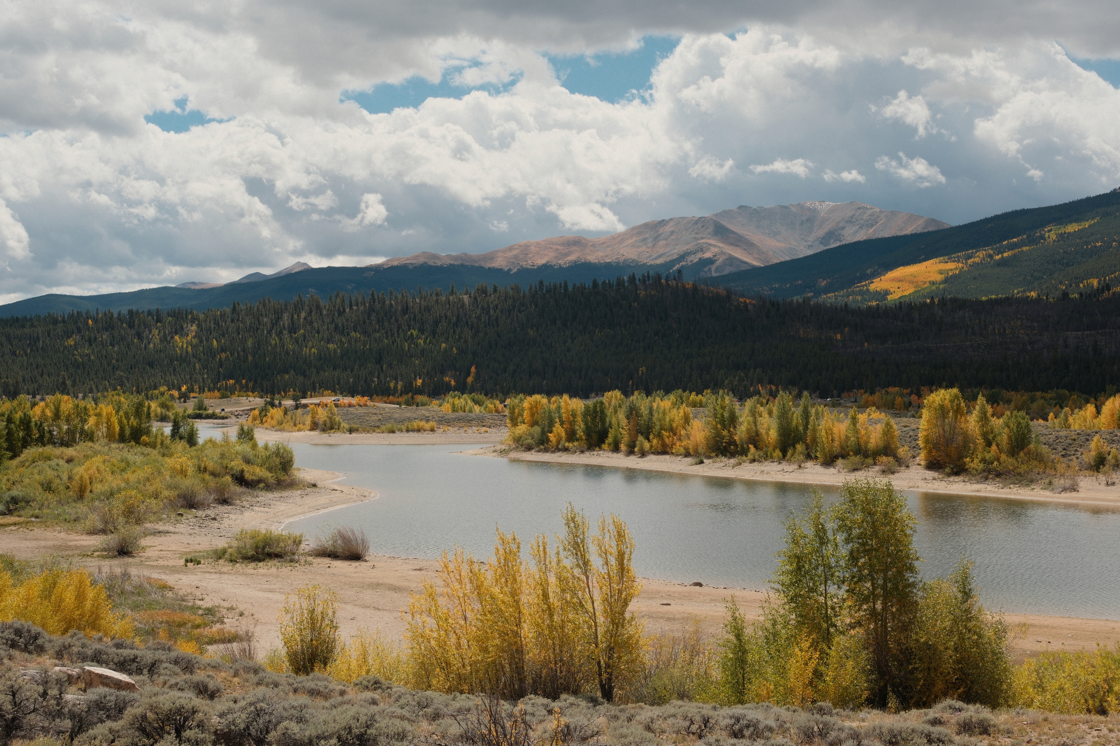 A mountain reservoir with a sandy shoreline, autumn aspens, and distant peaks under a cloudy sky