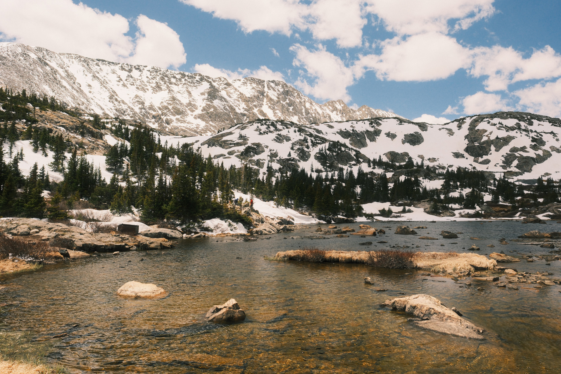 A clear alpine lake scattered with rocks below snow-covered peaks and scattered pines